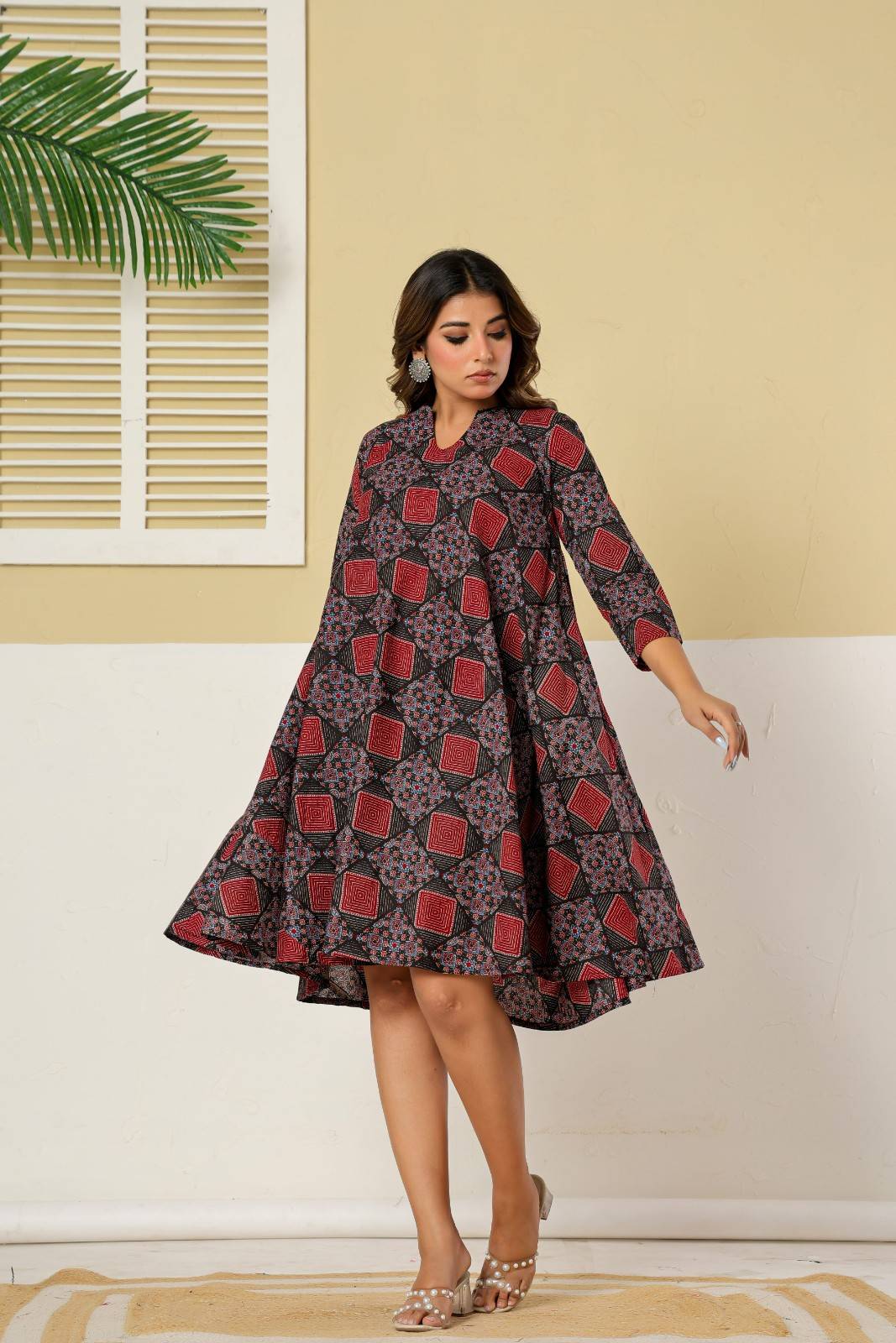 Woman wearing a patterned dress standing in a room with a plant and window blinds - YAVASTRAH