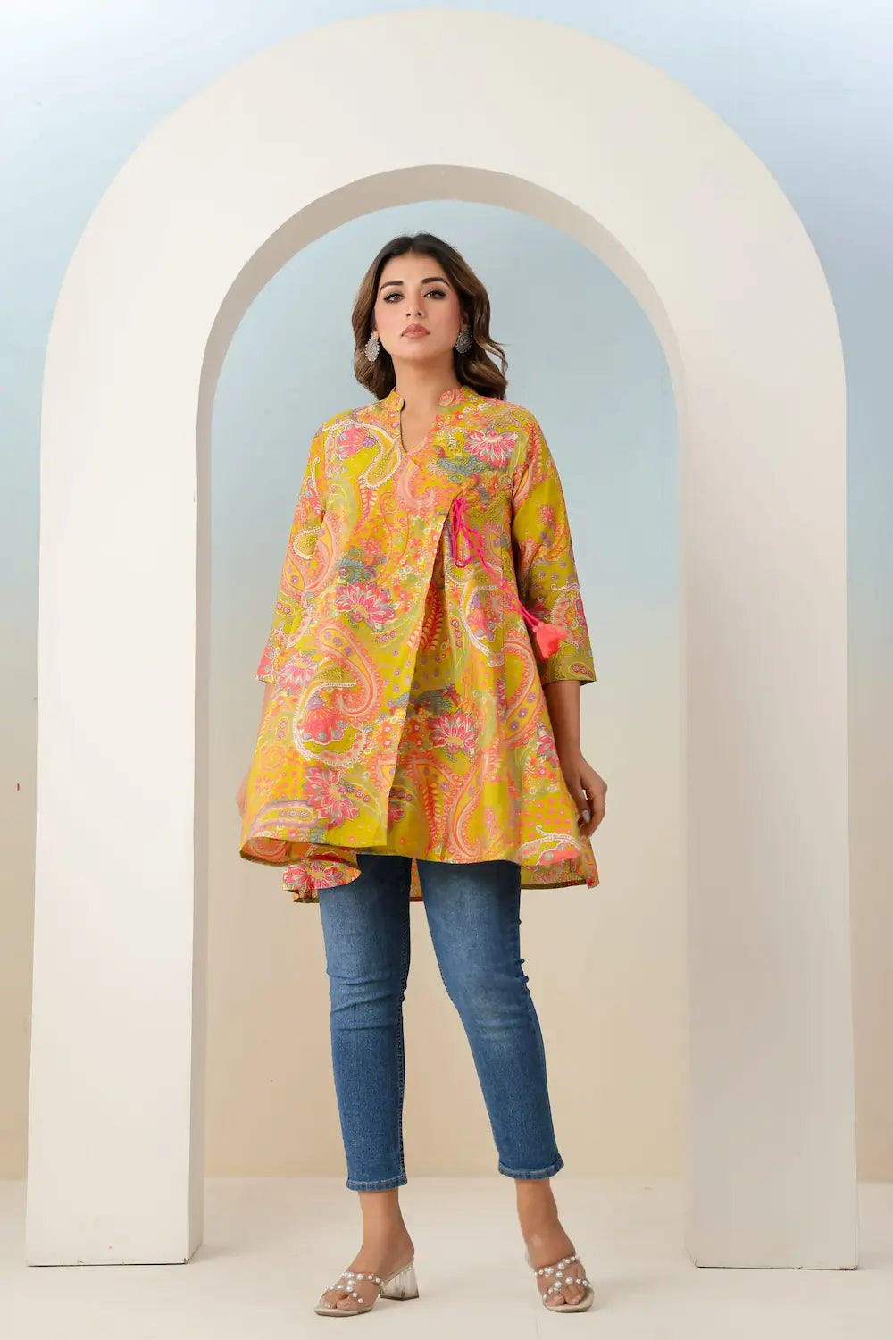 Woman wearing a colorful floral jacket and jeans standing in front of a white archway.