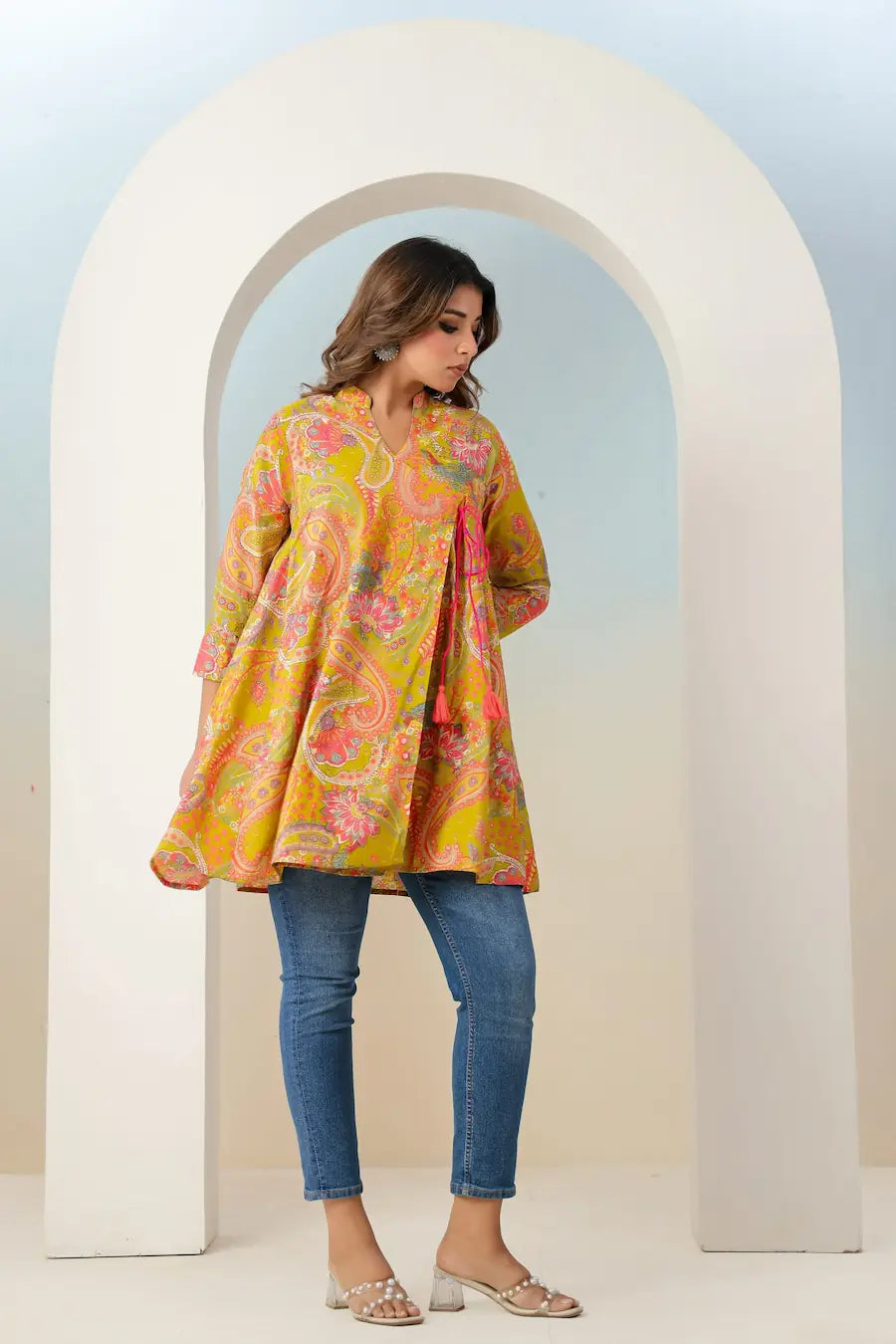 Woman wearing a colorful floral dress and jeans standing in front of a white archway.