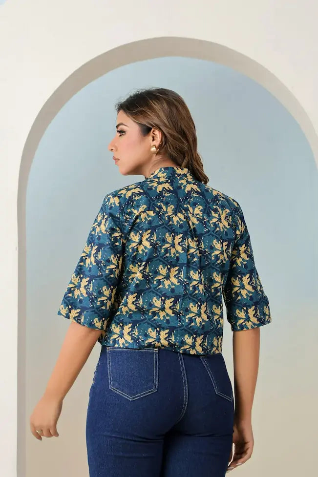 Woman wearing a blue floral blouse and dark jeans standing in front of an archway.