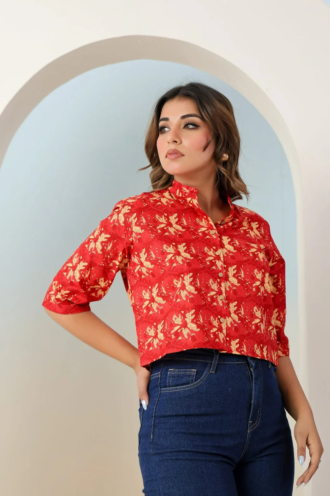 Woman wearing a red floral blouse and blue jeans standing in front of an archway - YAVASTRAH
