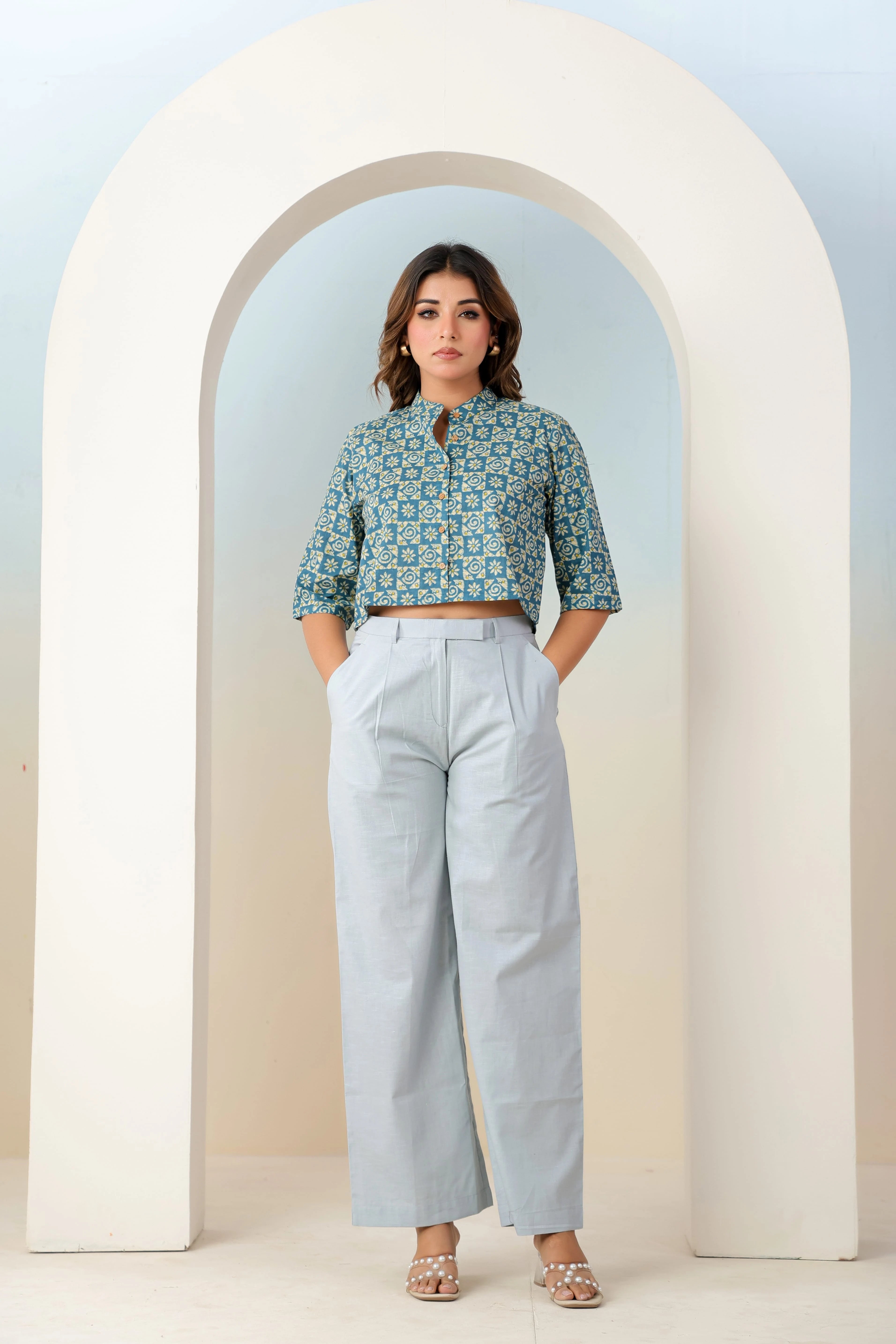 Woman wearing a patterned blouse and light gray pants standing in front of a white archway.