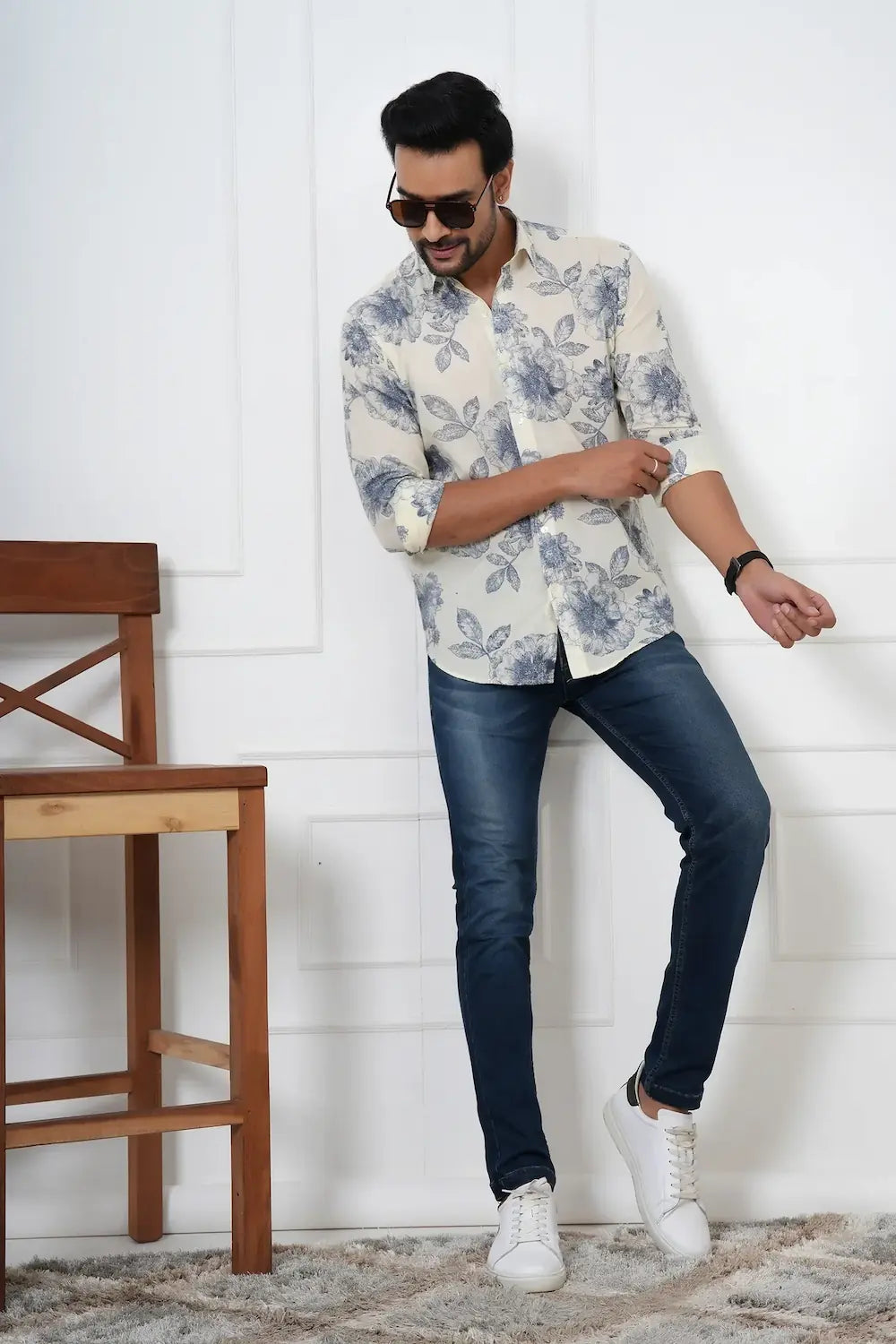Man wearing a floral shirt and dark jeans standing against a white wall.
