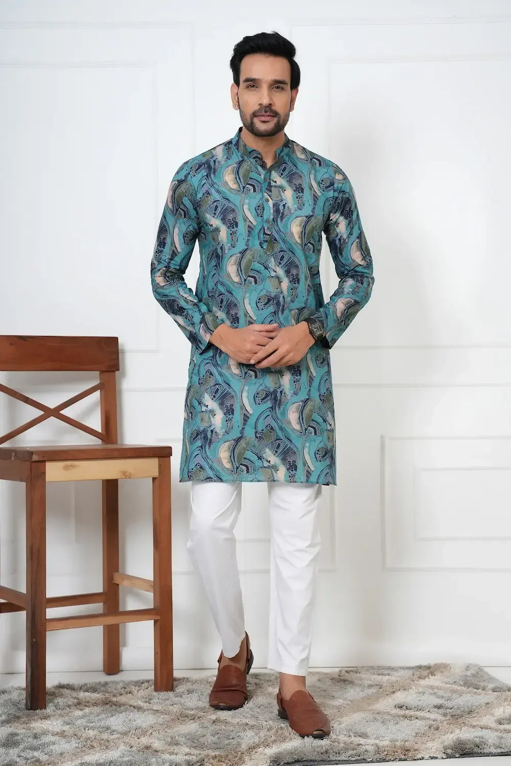 Man wearing a blue patterned kurta with white pants in a room with a wooden chair and white wall.