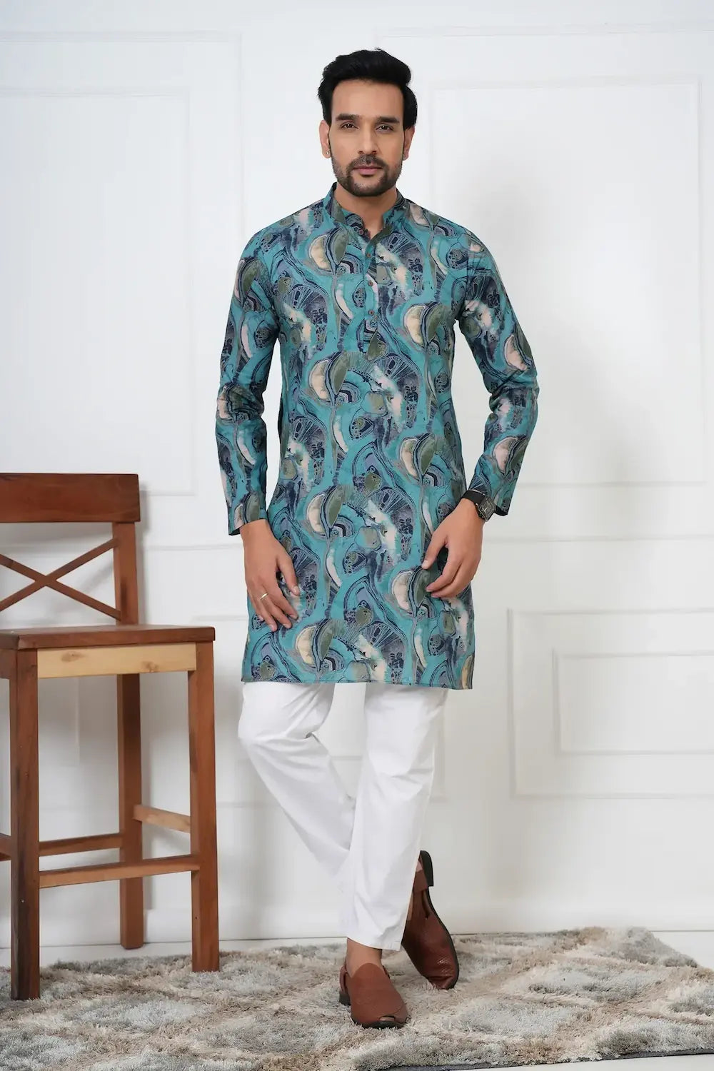 Man wearing a blue patterned kurta with white pants standing in a room with a wooden chair and white wall.