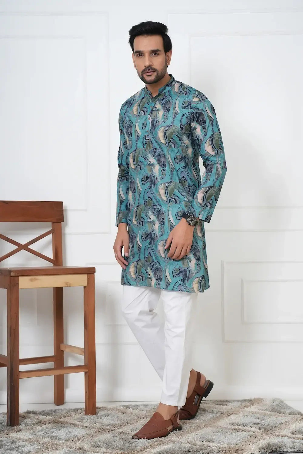 Man wearing a blue patterned kurta with white pants in a room with wooden chair and white wall.