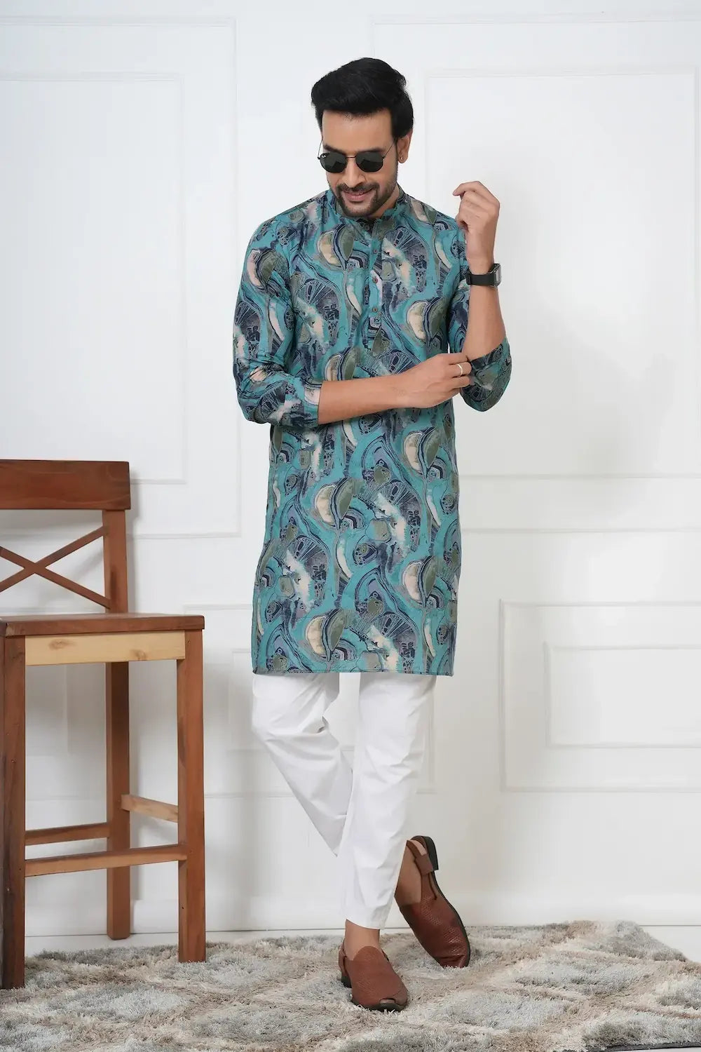 Man wearing a blue patterned kurta with white pants, standing in a room with a wooden chair and white wall.