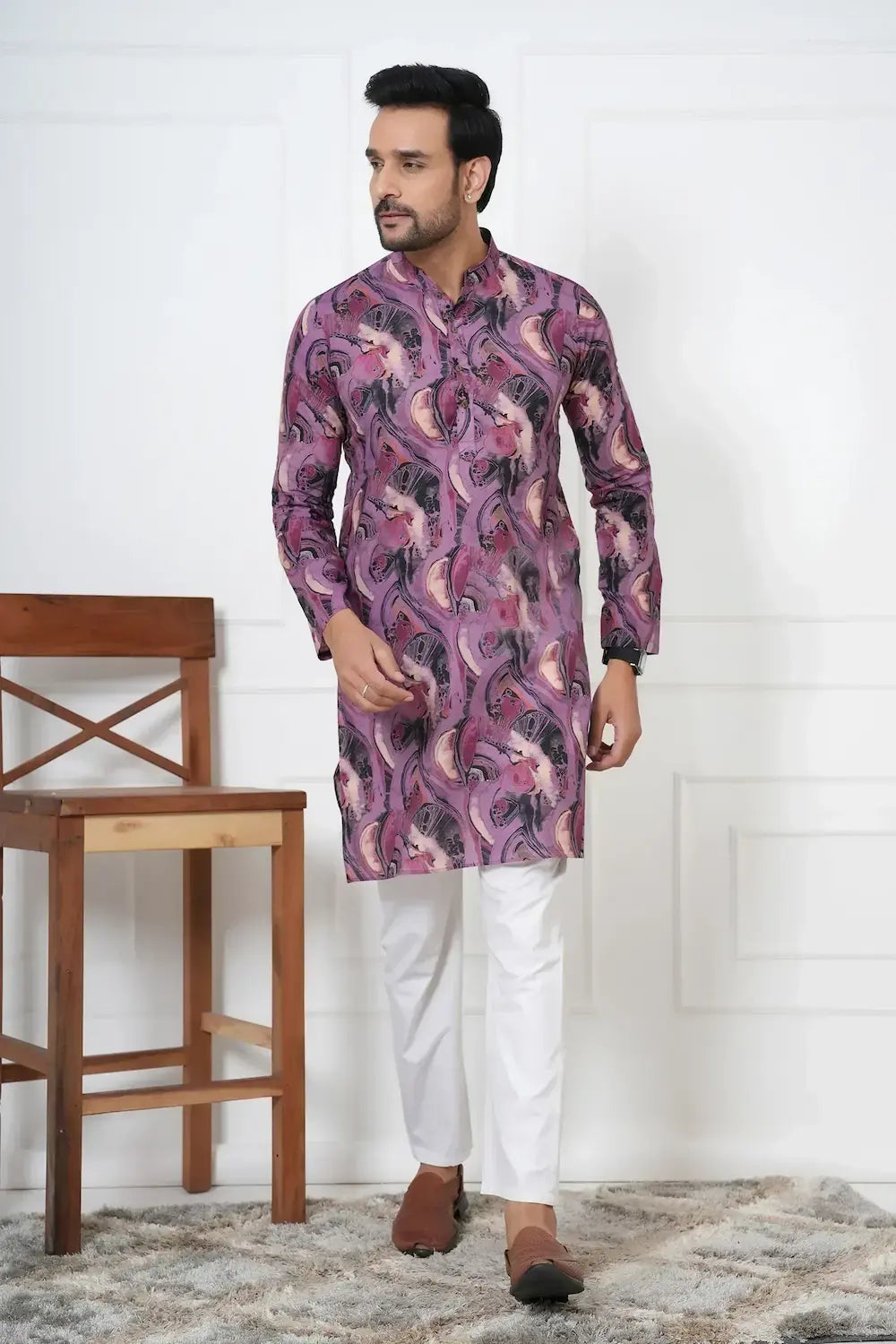 Man wearing a patterned kurta with white pants standing in a room with a wooden chair and white wall.