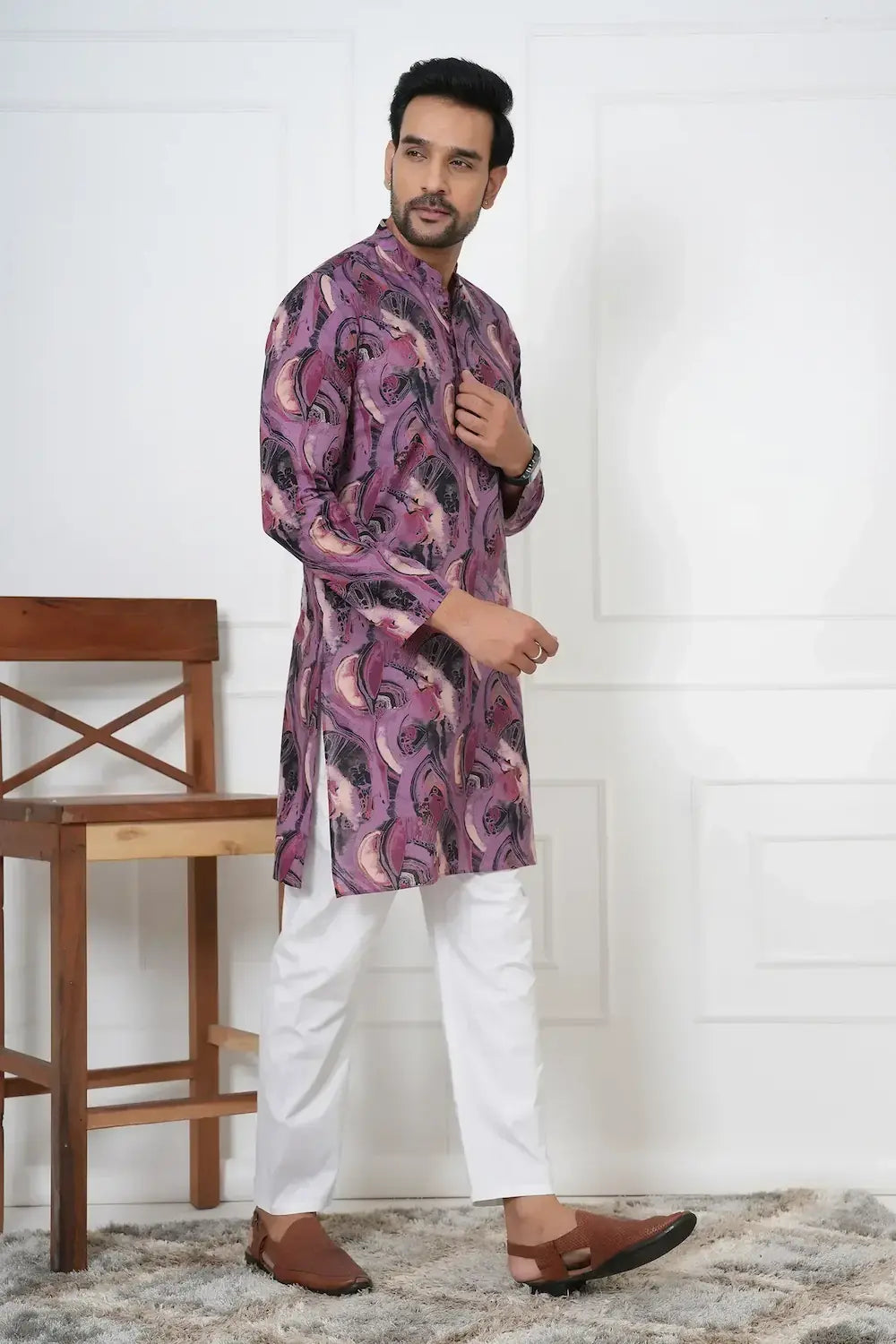 Man wearing a patterned kurta with white pants standing in a room with a wooden chair and white wall.
